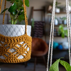 May include: A white ceramic planter with a green plant inside is hanging from a macrame plant hanger made of natural colored cotton rope. The plant hanger is crocheted with a mustard yellow yarn and has a geometric pattern.