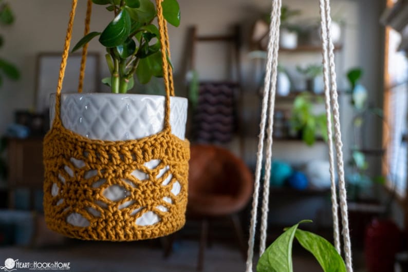 May include: A white ceramic planter with a green plant inside is hanging from a macrame plant hanger made of natural colored cotton rope. The plant hanger is crocheted with a mustard yellow yarn and has a geometric pattern.