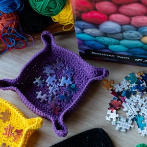 Può includere: Primo piano di un vassoio all'uncinetto viola con pezzi di puzzle. Sono visibili anche un vassoio all'uncinetto giallo e un oggetto all'uncinetto nero. Filato colorato e una scatola di puzzle "Rainbow Yarn" sullo sfondo.