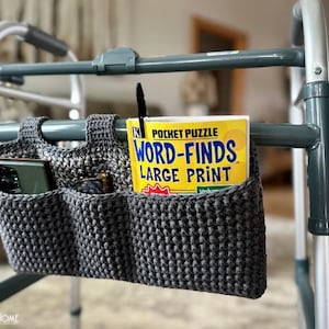 Op de afbeelding: Een grijze gehaakte tas met drie vakken hangt aan een rollator. De tas bevat een telefoon, een pen en een boek met de titel "Word-Finds Large Print Pocket Puzzle".