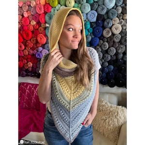 May include: A crocheted hooded scarf in shades of yellow, blue, and gray. The scarf is worn by a person with long brown hair. The background is a wall of colorful yarn balls.
