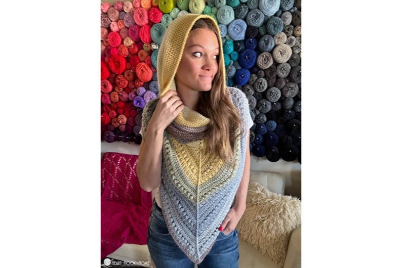 May include: A crocheted hooded scarf in shades of yellow, blue, and gray. The scarf is worn by a person with long brown hair. The background is a wall of colorful yarn balls.