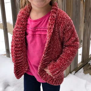 May include: A red crocheted cardigan with a shawl collar. The cardigan is made with a fuzzy yarn and has a slightly textured look.