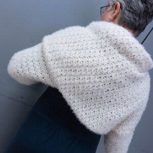 May include: A white crocheted shrug with a fuzzy texture. The shrug has long sleeves and a cropped length.