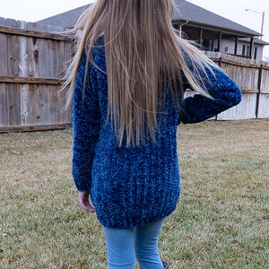 May include: A blue fuzzy sweater with long sleeves and a long length. The sweater is worn by a person with long brown hair. The person is wearing blue jeans and brown boots.