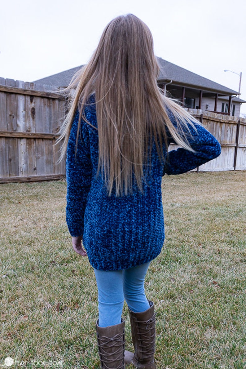 May include: A blue fuzzy sweater with long sleeves and a long length. The sweater is worn by a person with long brown hair. The person is wearing blue jeans and brown boots.