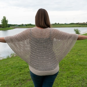 May include: A woman wearing a beige crochet top with a loose, open-weave design. The top has a wide, flowing neckline and long, wide sleeves. The woman is standing outdoors with her arms outstretched, facing away from the camera.