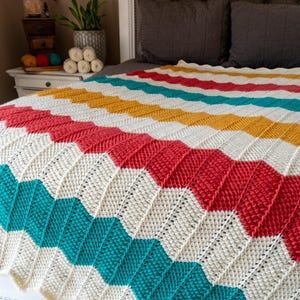 May include: A colorful, handmade crochet blanket with a chevron pattern. The blanket features stripes of white, teal, red, yellow, and turquoise. The blanket is displayed on a bed, showcasing its vibrant design and texture.