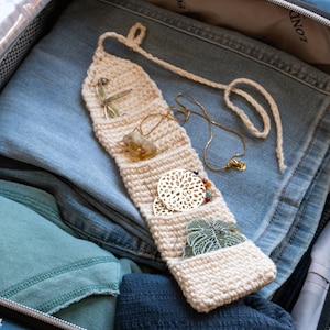 Puede incluir: Un organizador de joyas de ganchillo color crema con múltiples bolsillos, que contiene varios collares, aretes y objetos decorativos. El organizador está dentro de una maleta con jeans azules y otras prendas.