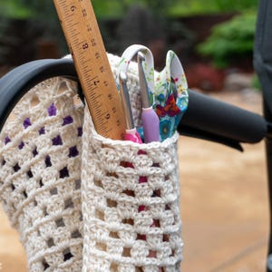 Puede incluir: Una bolsa de crochet blanca con detalles morados cuelga de una silla de metal negro. La bolsa está llena de una regla, un par de agujas de crochet y un par de tijeras.