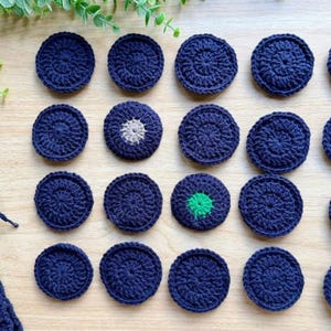 May include: Twenty circular navy blue crochet pieces arranged in a grid. One piece features a small light gray center, and another features a bright green center. A partially visible navy blue crocheted item with a long yarn tail is at the bottom left.