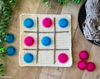 May include: A square, off-white crocheted tic-tac-toe board featuring gray stitched grid lines. The board contains five translucent blue and four translucent pink circular plastic game pieces, with additional loose pieces placed nearby.