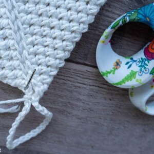 May include: Close-up of a white crocheted piece with a needle and white yarn. The yarn is being threaded through the crochet stitches. A pair of floral patterned scissors are in the background.