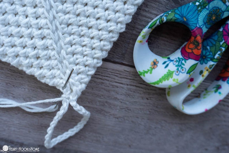 May include: Close-up of a white crocheted piece with a needle and white yarn. The yarn is being threaded through the crochet stitches. A pair of floral patterned scissors are in the background.