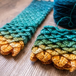 May include: A close-up of a crocheted scarf with a dragon scale pattern in shades of green, yellow, and orange. The scarf is made with a gradient yarn and is lying on a wooden surface.