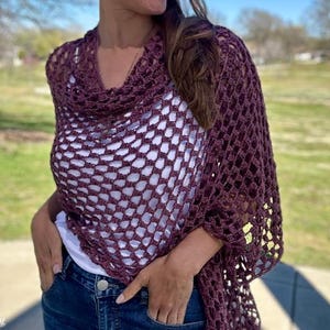 Puede incluir: Un chal de ganchillo de color morado oscuro con un diseño de encaje de trabajo abierto. El chal está drapeado sobre los hombros de una persona y se lleva con una camisa blanca y unos vaqueros azules.