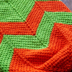 May include: A handmade crochet blanket in a vibrant color scheme of green and orange. The blanket features a geometric design with alternating bands of the two colors, creating a textured and visually appealing pattern. The stitches are tightly woven, suggesting a durable and cozy item.