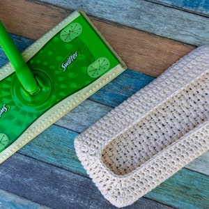 May include: A white crocheted storage basket for Swiffer mop pads. The basket is lying on a wooden surface with a green Swiffer mop in the background.