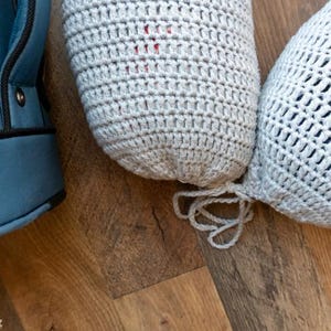 Puede incluir: Primer plano de dos fundas de botella de ganchillo en gris claro, una con un tejido m&aacute;s apretado y la otra con un dise&ntilde;o m&aacute;s abierto y calado. Una bolsa azul es parcialmente visible a la izquierda. Los art&iacute;culos est&aacute;n sobre una superficie de madera.