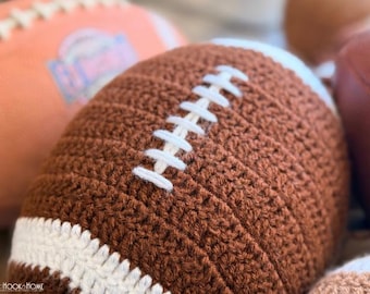 May include: A brown crocheted football featuring white yarn laces and white stripes at the ends. The textured yarn surface mimics the appearance of a traditional football. A small logo with the text Heart Hook Home is visible in the corner.