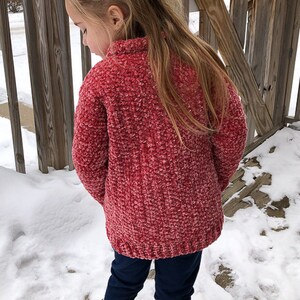 May include: A young girl wearing a red fuzzy sweater and blue jeans stands in the snow. She is wearing a pink hair tie in her hair.