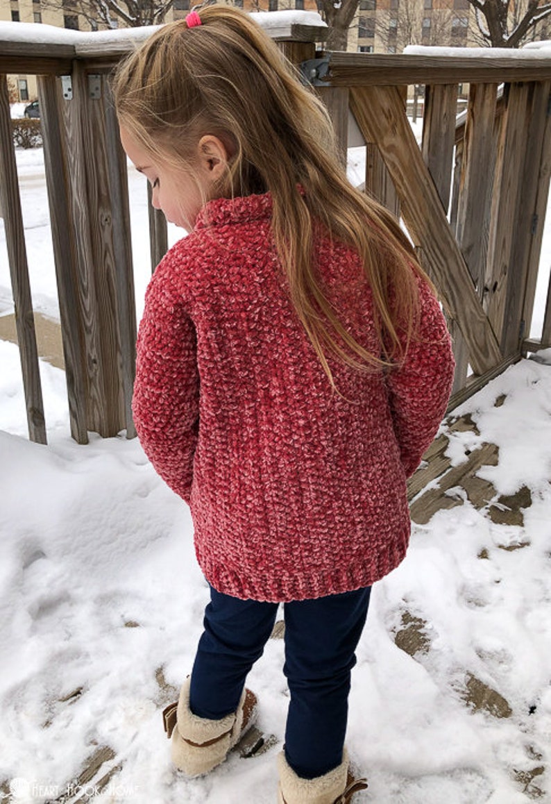 May include: A young girl wearing a red fuzzy sweater and blue jeans stands in the snow. She is wearing a pink hair tie in her hair.