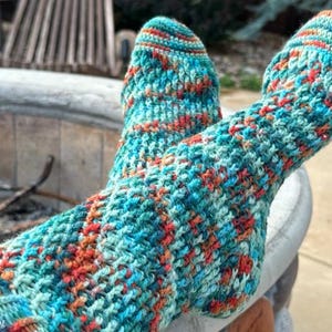 Könnte beinhalten: Nahaufnahme von zwei Füßen mit handgestrickten Socken. Die Socken sind eine Mischung aus türkis, orange und roter Wolle mit einem strukturierten, gewebten Muster. Die Füße ruhen auf einer Betonoberfläche mit verschwommenem Hintergrund im Freien.