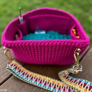 May include: A vibrant fuchsia crochet bag with a colorful patterned strap. The bag has interior pockets and a teal lining. A crochet hook is visible inside the bag. Gold-colored hardware connects the strap to the bag.