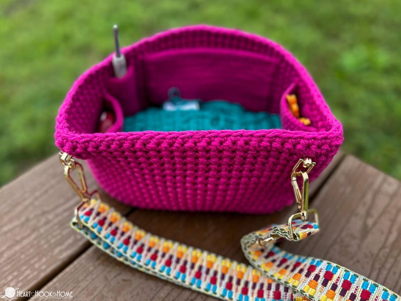 May include: A vibrant fuchsia crochet bag with a colorful patterned strap. The bag has interior pockets and a teal lining. A crochet hook is visible inside the bag. Gold-colored hardware connects the strap to the bag.