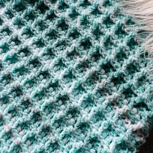 May include: Close-up of a hand-crocheted blanket with a waffle stitch pattern. The blanket is a blend of teal, light blue, and white yarn, creating a textured appearance. The edge of the blanket has a fringe of white faux fur.