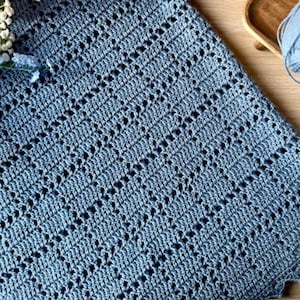 May include: A handmade, blue crochet blanket with a scalloped edge. The blanket features a delicate openwork design and is made of soft yarn. A ball of blue yarn and flowers are in the background.