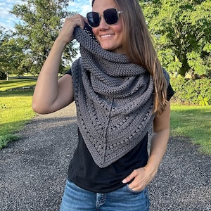 May include: A woman wearing a gray triangular shawl with a textured pattern. She is wearing a black shirt and blue jeans. The shawl is draped over her shoulders and she is holding it up with her right hand.