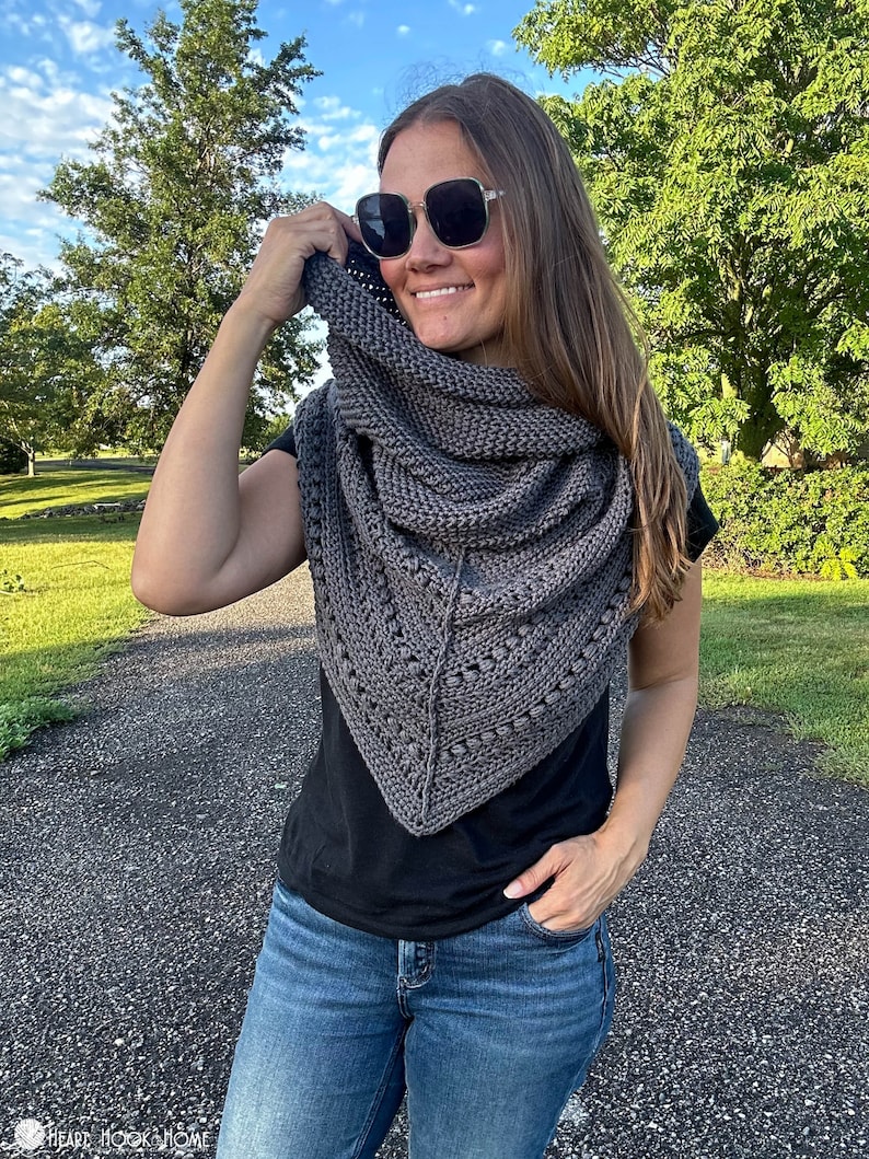 May include: A woman wearing a gray triangular shawl with a textured pattern. She is wearing a black shirt and blue jeans. The shawl is draped over her shoulders and she is holding it up with her right hand.