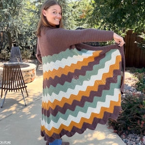 May include: A long-sleeved crochet cardigan featuring a chevron pattern in alternating horizontal stripes of brown, sage green, mustard yellow, and cream. The garment has a solid brown upper section and long sleeves, with a flared, wavy hemline.