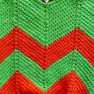 May include: A handmade crochet item with a vibrant chevron pattern. The design features alternating bands of bright green and orange, creating a bold and eye-catching visual effect. The texture of the crochet stitch is clearly visible.