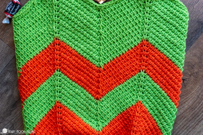 May include: A handmade crochet item with a vibrant chevron pattern. The design features alternating bands of bright green and orange, creating a bold and eye-catching visual effect. The texture of the crochet stitch is clearly visible.