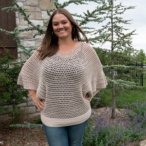 May include: A woman wearing a beige crochet top with a loose, open-weave design. The top has a wide, rounded neckline and long, flowing sleeves. The woman is standing in front of a stone wall and green foliage.