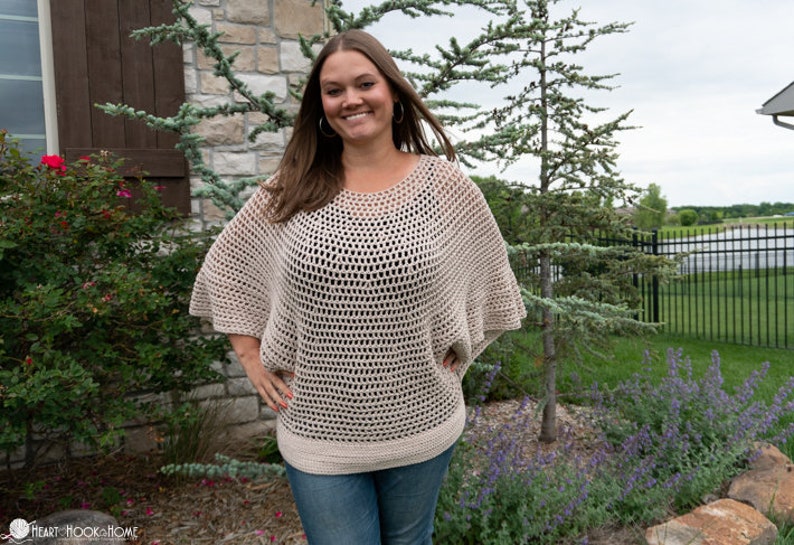 May include: A woman wearing a beige crochet top with a loose, open-weave design. The top has a wide, rounded neckline and long, flowing sleeves. The woman is standing in front of a stone wall and green foliage.
