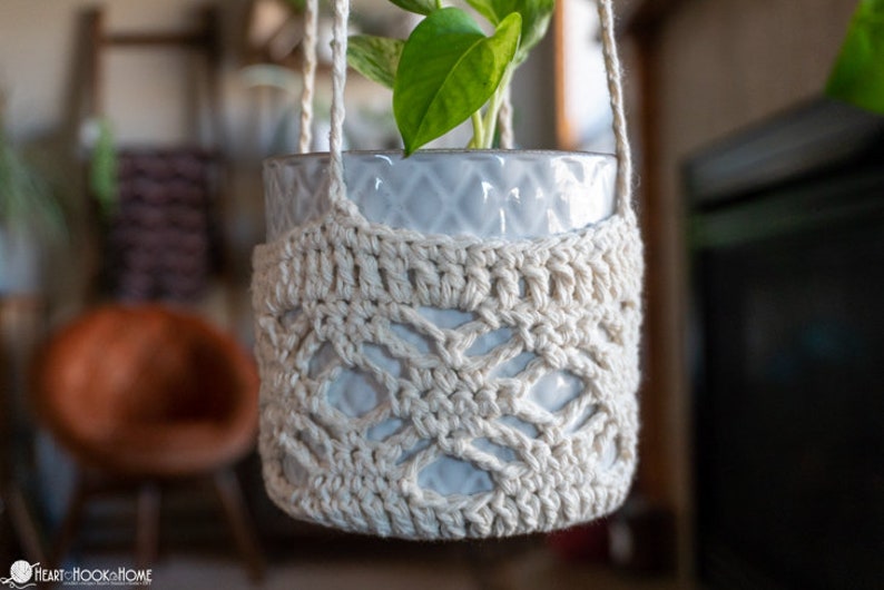 May include: A white crocheted plant hanger with a diamond pattern, holding a white ceramic pot with a green plant.