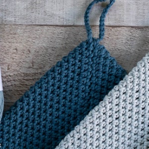 May include: Three crocheted dishcloths in shades of blue, grey, and beige. The dishcloths have a textured, waffle-like pattern and are made with 100% cotton yarn. The yarn is labelled "Rainbow 100% Cotton" and "Hobbii".