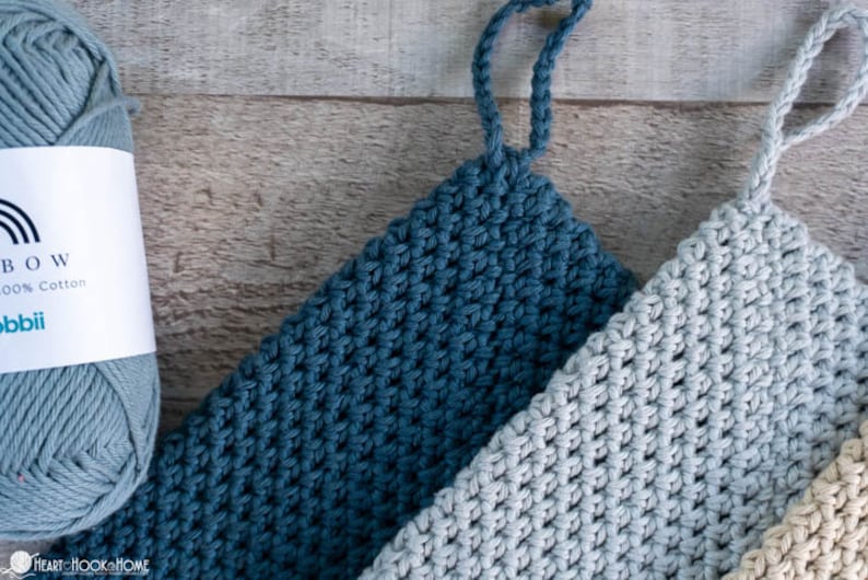May include: Three crocheted dishcloths in shades of blue, grey, and beige. The dishcloths have a textured, waffle-like pattern and are made with 100% cotton yarn. The yarn is labelled "Rainbow 100% Cotton" and "Hobbii".