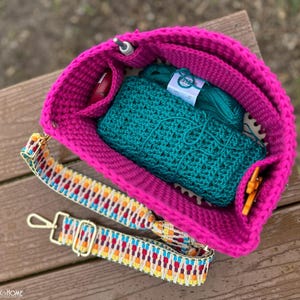 May include: A bright pink crochet bag with a colorful strap, holding teal yarn and a teal crochet project. The bag has pockets for small tools. The strap has a geometric pattern with gold-colored hardware.