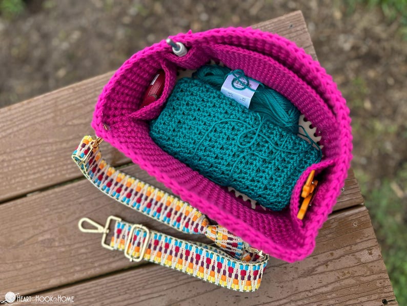 May include: A bright pink crochet bag with a colorful strap, holding teal yarn and a teal crochet project. The bag has pockets for small tools. The strap has a geometric pattern with gold-colored hardware.