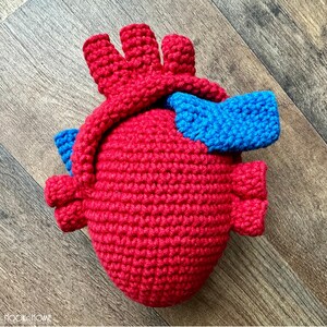May include: A crocheted anatomical heart plush toy featuring a vibrant red main body with textured yarn stitches. The heart includes prominent blue crocheted vessels attached to the top and sides, mimicking the structure of a human heart.