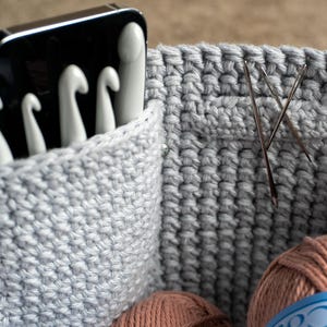May include: Close-up of a gray crocheted basket with white crochet hooks in a pocket. Two needles are inserted into the basket's side. Two balls of yarn are visible at the bottom of the basket. The basket is designed for crochet supplies.