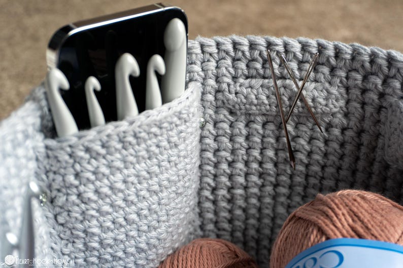 May include: Close-up of a gray crocheted basket with white crochet hooks in a pocket. Two needles are inserted into the basket's side. Two balls of yarn are visible at the bottom of the basket. The basket is designed for crochet supplies.