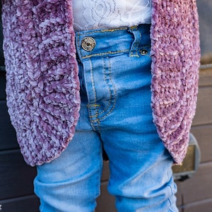 May include: A close-up of a child wearing a purple fuzzy cardigan over a pair of blue jeans. The cardigan is made of a soft, fluffy material and has a slightly textured look. The jeans are a light blue wash and have a classic five-pocket style.