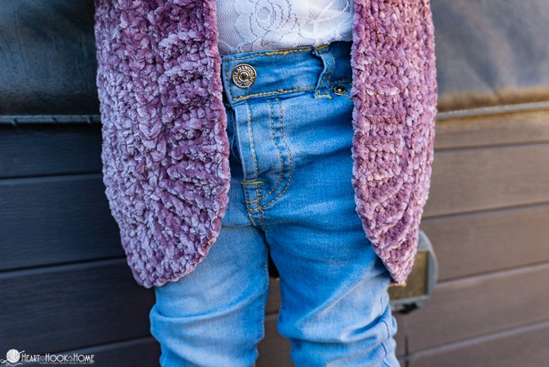 May include: A close-up of a child wearing a purple fuzzy cardigan over a pair of blue jeans. The cardigan is made of a soft, fluffy material and has a slightly textured look. The jeans are a light blue wash and have a classic five-pocket style.
