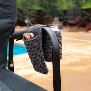 May include: Two gray crocheted can cozies are attached to the armrest of a black folding chair. The cozies are holding two aluminum cans.