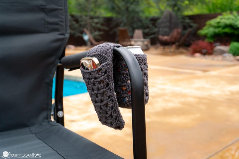 May include: Two gray crocheted can cozies are attached to the armrest of a black folding chair. The cozies are holding two aluminum cans.